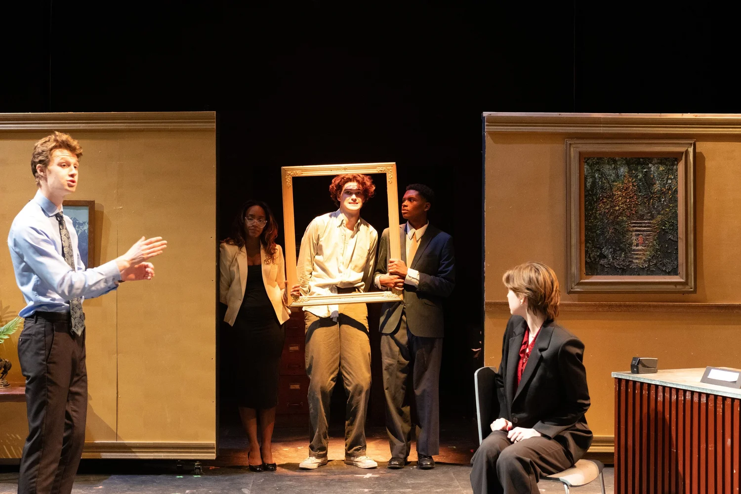 Photo of an actor inside of a frame held by two other actors in the background while two more actors argue in the foreground.